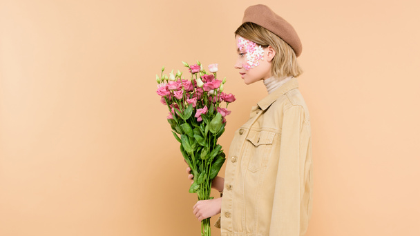 side view of stylish woman with flowers on face holding bouquet isolated on beige  - Foto, afbeelding