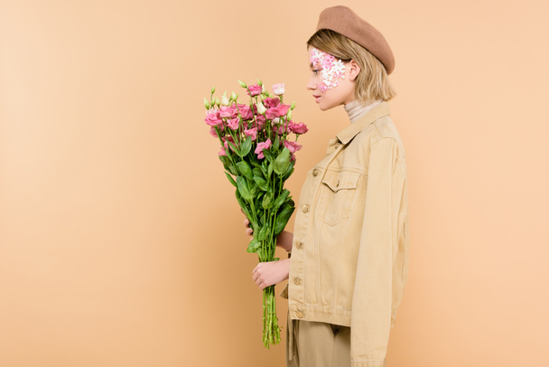 profile of stylish woman with flowers on face holding bouquet isolated on beige  - Foto, afbeelding