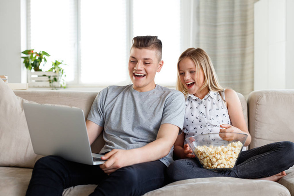 Teenage boy and his sister sitting on sofa at home and watching funny movie on laptop, eating popcorn.  - Zdjęcie, obraz