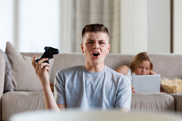 Brother and sister spending time at home, sitting on sofa in living room. Boy playing video game and girl using digital tablet.  - Foto, Bild