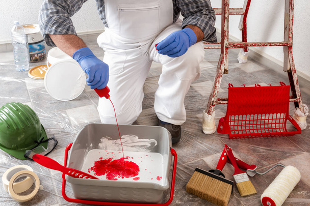Pintor de casa caucásico trabajador en overoles de trabajo blanco, vierte tinte rojo en el recipiente con pintura blanca. Industria de construcción
. - Foto, imagen