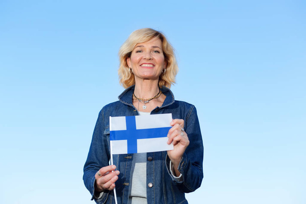 glückliche reife Frau mit finnischer Flagge vor dem Hintergrund eines blauen Himmels. - Foto, Bild