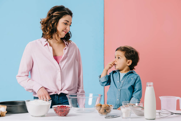 madre feliz con adorable hijito por mesa de cocina con ingredientes y utensilios de cocina sobre fondo bicolor
 - Foto, imagen