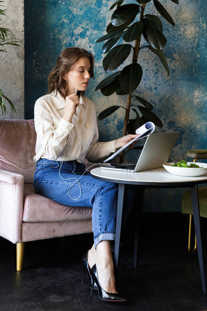 Photo of pretty beautiful young woman sitting in cafe indoors using laptop holding documents talking by mobile phone. - Foto, Bild