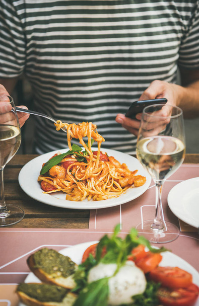 Italiaans diner in bistrot met caprese salade met mozzarella, tomaten, basilicum, toast van pesto, spaghetti pasta met garnalen, witte wijn. Man eten pasta met smartphone. Heerlijke Italiaanse gerechten - Foto, afbeelding