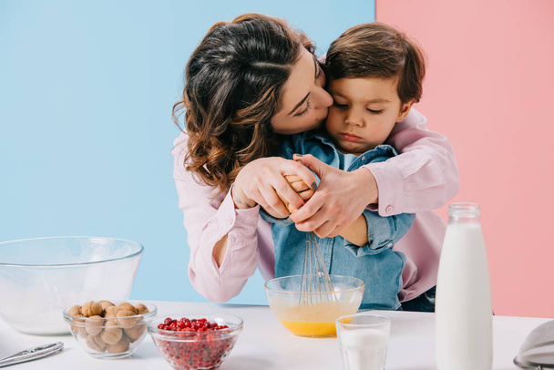 mamma baciare adorabile piccolo figlio mentre montare le uova con palloncino frusta insieme su sfondo bicolore
 - Foto, immagini