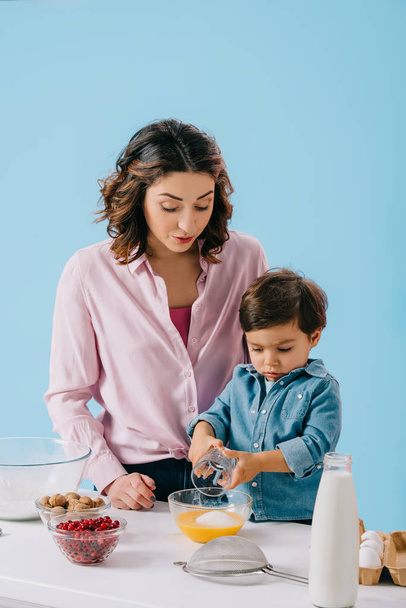 bonito pequeno menino adicionando açúcar para chicoteado ovos enquanto mamãe olhando para ele isolado no azul
 - Foto, Imagem