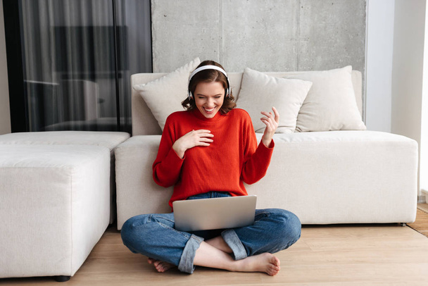 Happy young casually dressed woman sitting on a floor at home, using laptop computer, celebrating success - Фото, изображение