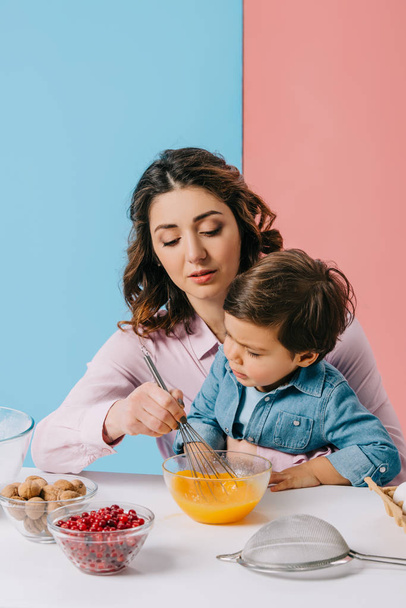 moeder zit met zoontje door keukentafel en zweepslagen eitjes in kom op bicolor achtergrond - Foto, afbeelding