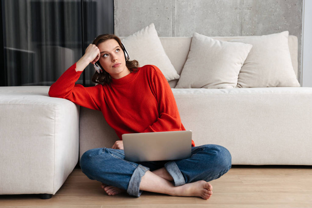 Müde junge, lässig gekleidete Frau sitzt zu Hause auf einem Fußboden und lernt mit Laptop - Foto, Bild