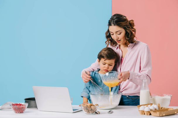 mother adding whipped eggs to bowl while cooking together with little son on bicolor background - Photo, Image