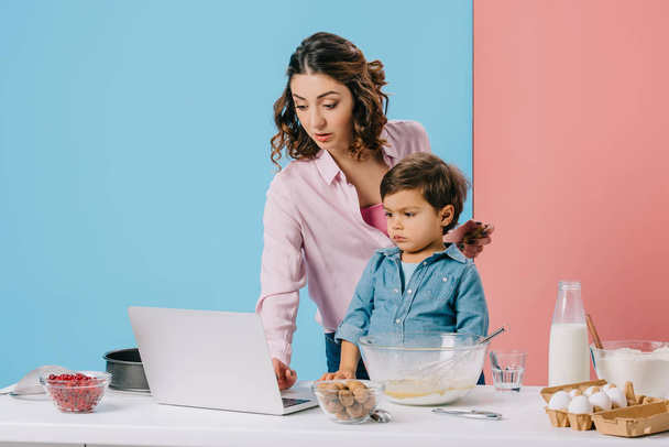 madre usando escritorio mientras cocina junto con su hijo pequeño sobre fondo bicolor
 - Foto, Imagen