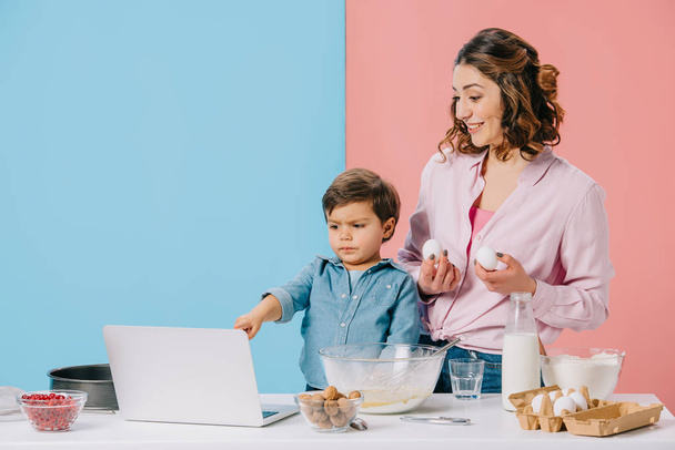 bambino che punta al display del computer portatile durante la cottura con la madre su sfondo bicolore
  - Foto, immagini
