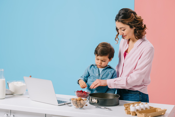 Lächelnde Mutter mit kleinem Sohn, die beim gemeinsamen Kochen am Küchentisch mit Kochzutaten und Laptop auf zweifarbigem Hintergrund Preiselbeeren zur Backform hinzufügt  - Foto, Bild