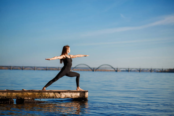 Young woman practicing yoga exercise at quiet wooden pier with city background. Sport and recreation in city rush. - Photo, Image