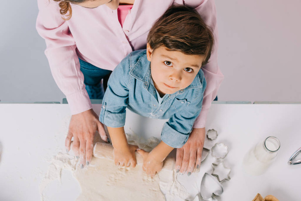 top view of cute little boy rolling out dough with mother and looking at camera on light background - Foto, Bild