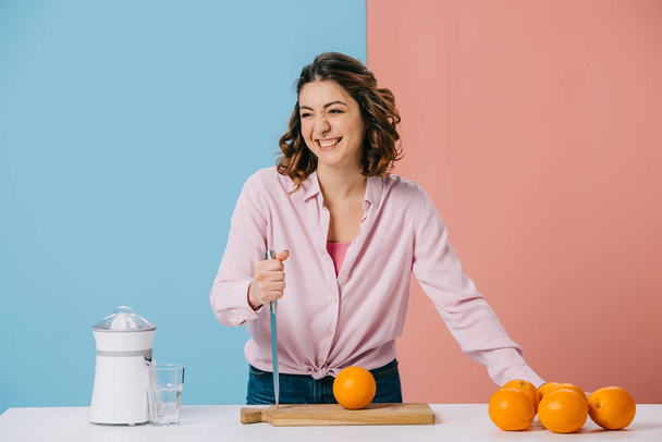 魅力的な女性は二色の背景の上に新鮮なオレンジを台所のテーブルで立ちながらナイフを所持  - 写真・画像