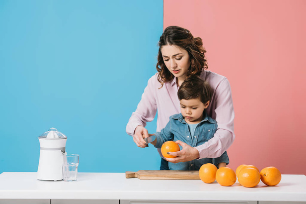 χαριτωμένο μικρό αγόρι που βλέπουν πορτοκάλι στο χέρι μητέρες ενώ στέκεται με τραπέζι κουζίνας με πορτοκάλια σε δίχρωμο φόντο  - Φωτογραφία, εικόνα