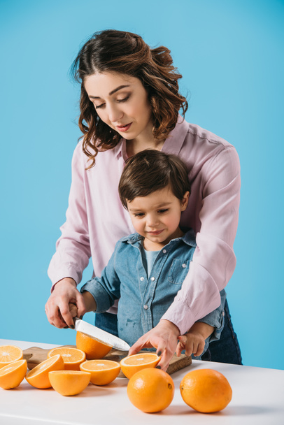 mãe bonita com pequeno filho bonito cortando laranjas na placa de corte de madeira juntos isolados em azul
 - Foto, Imagem