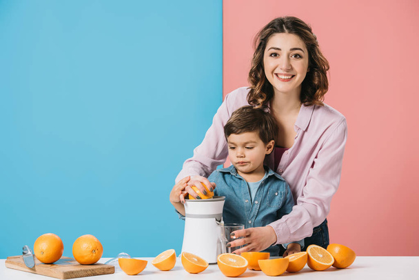 happy mother squeezing fresh orange juice together with cute little son on bicolor background - Photo, Image