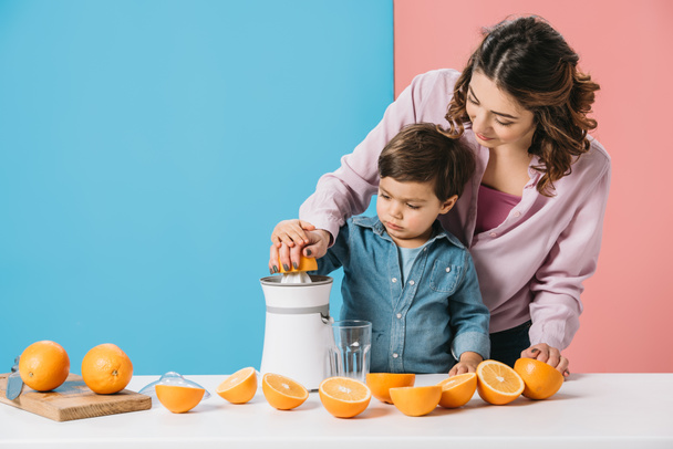 madre con lindo hijo pequeño exprimiendo jugo de naranja fresco juntos sobre fondo bicolor
 - Foto, Imagen