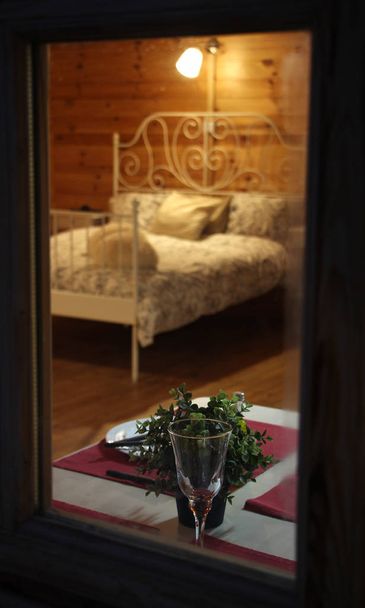 шпионский вид через окно деревянного загородного дома, постельное белье, накрытый стол для романтического ужина, бокал вина, тарелки и ночник, секретное рандеву
 - Фото, изображение