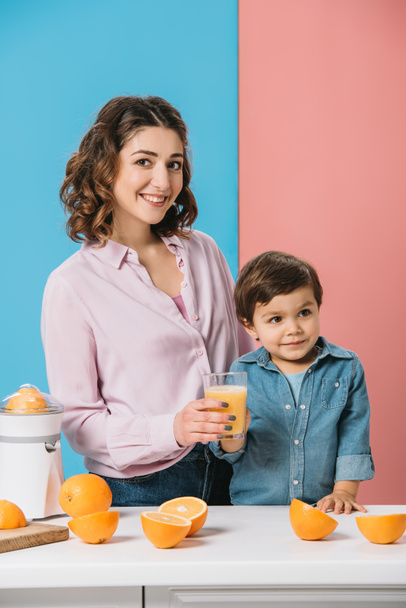 愛らしいに一杯の新鮮なオレンジ ジュースを与えること幸せな母笑顔二色の背景に幼い息子  - 写真・画像