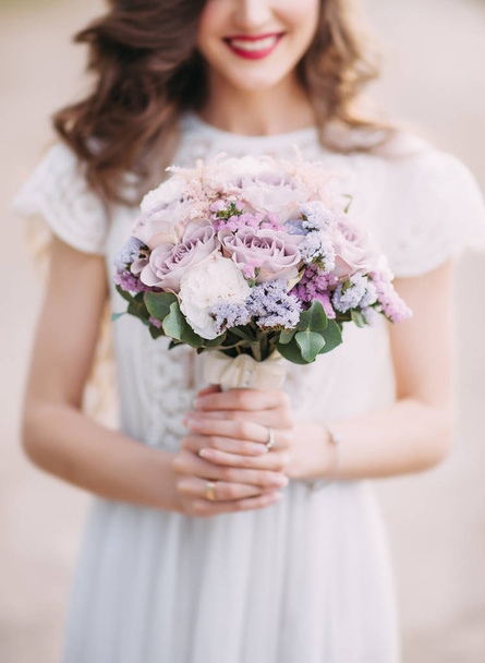 Wedding bouquet in white and violet colours, close up. Modest beautiful smiling bride with round purple wedding bouquet. White lace and silk wedding dress. Happy wedding concept - Фото, изображение