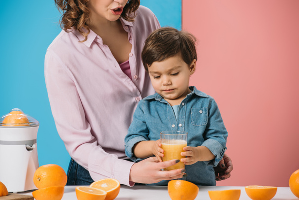 cute little boy taking glass full of fresh orange juice from mothers hand on bicolor background  - Photo, Image