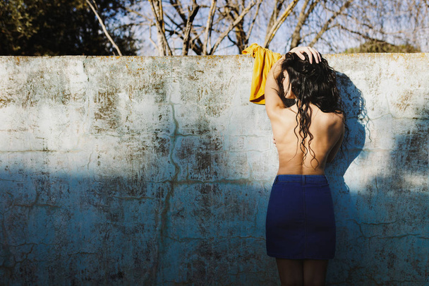 Beautiful brunette against the wall of an empty pool undressing with the sun illuminating her naked back - Photo, Image