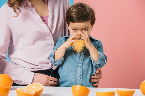 adorable little boy enjoying fresh orange juice while standing with mother by kitchen table on bicolor background  - Photo, Image