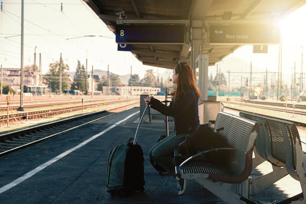 Młoda Ruda kobieta czeka następny pociąg na stacji z jej walizkę i plecak - Zdjęcie, obraz