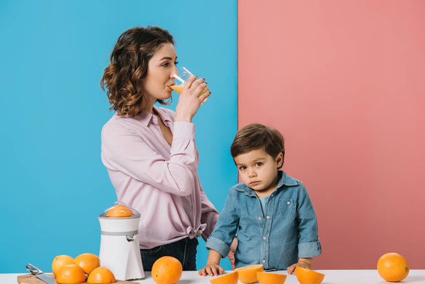 mother drinking fresh orange juice while standing with cute little son by table with oranges on bicolor background  - Photo, Image