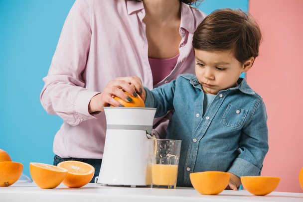 adorable little boy squeezing fresh orange juice on juicer together with mother on bicolor background  - Foto, imagen