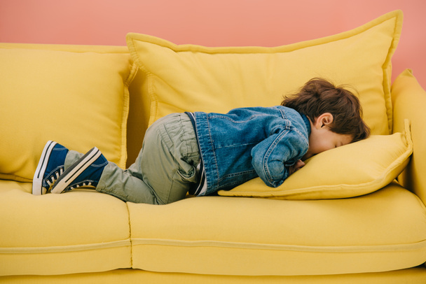 Niedlicher kleiner Junge liegt mit dem Gesicht nach unten auf bequemem gelben Sofa - Foto, Bild