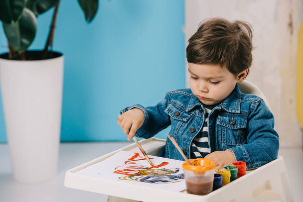 cute little boy painting with watercolor paints while sitting on highchair - Zdjęcie, obraz