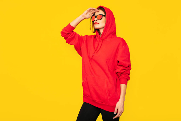 cool and tuff hipster girl with sunglasses posing in red hoodie on yellow background - Foto, Imagem