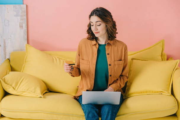 smiling attractive woman holding credit card while sitting on yellow sofa and using laptop - Foto, Imagem