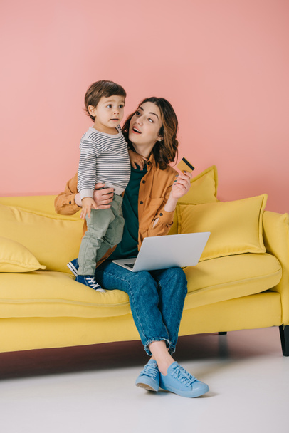 sonriente madre con portátil en las rodillas sosteniendo la tarjeta de crédito y abrazando a su pequeño hijo de pie cerca en el sofá amarillo
 - Foto, imagen