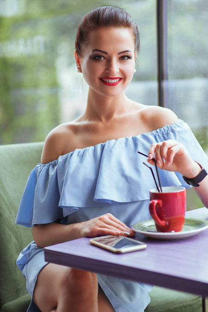 Mosolygós boldog üzletasszony, mozgatható telefon és piros csésze kávét a kávézóban. Csupasz vállát a kávézóban kék ruha gyönyörű nő portréja. Gyönyörű női barna modell - Fotó, kép