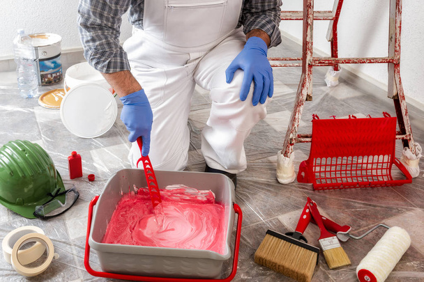 Pintor de casa caucásico trabajador en mono de trabajo blanco, mezcla la pintura en el recipiente. Industria de construcción
. - Foto, imagen