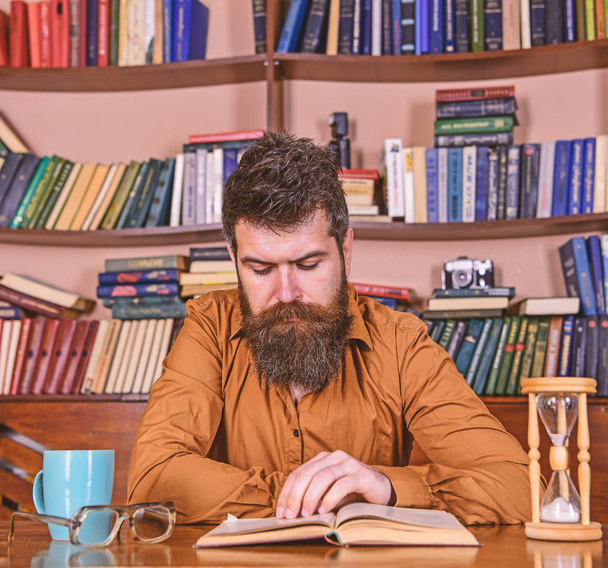Man on concentrated face reading book, studying, bookshelves on background. Self education concept. Teacher or student with beard studying in library. Man sits at table with mug and hourglass - Fotó, kép