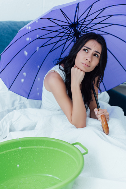 Chica triste con paraguas mirando el agua que gotea del techo en el dormitorio
 - Foto, Imagen