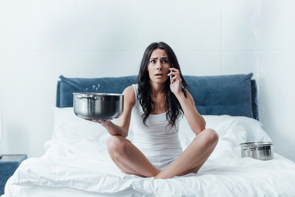 Mujer conmocionada hablando en el teléfono inteligente y sosteniendo la olla bajo el techo con fugas en el dormitorio
 - Foto, Imagen
