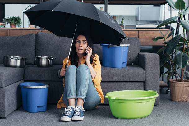 Girl in jeans sitting in living room with umbrella and talking on smartphone - Photo, Image