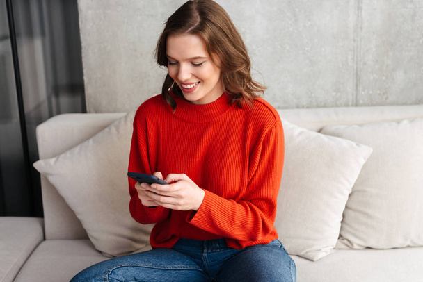 Cheerful young casually dressed woman sitting on a couch at home, holding mobile phone - Фото, изображение