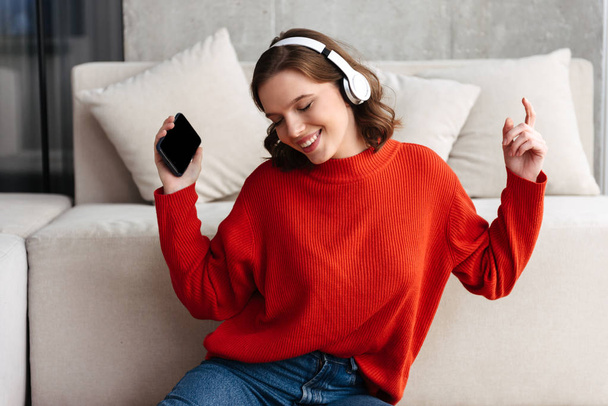 Iloinen nuori rennosti pukeutunut nainen istuu lattialla kotona, kuunnellen musiikkia kuulokkeilla
 - Valokuva, kuva