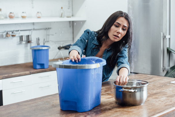 Jovem estressada usando baldes e panela durante vazamento na cozinha
 - Foto, Imagem