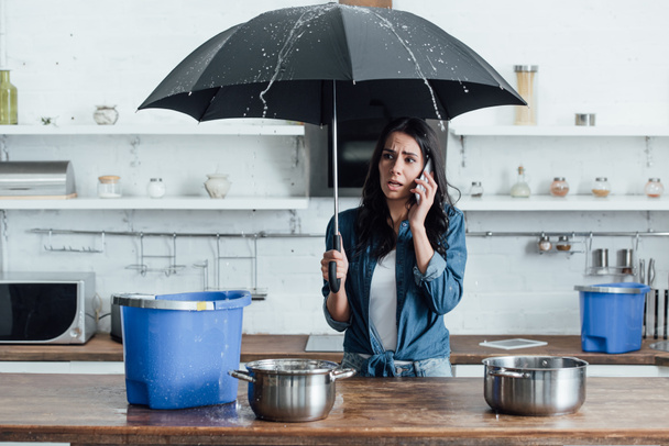 Upset woman standing under umbrella in kitchen and calling plumber - Photo, Image