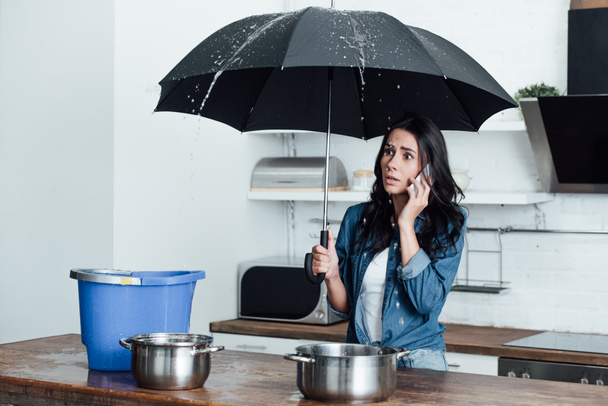 Shocked woman with umbrella dealing with water damage in kitchen and talking on smartphone - Photo, Image